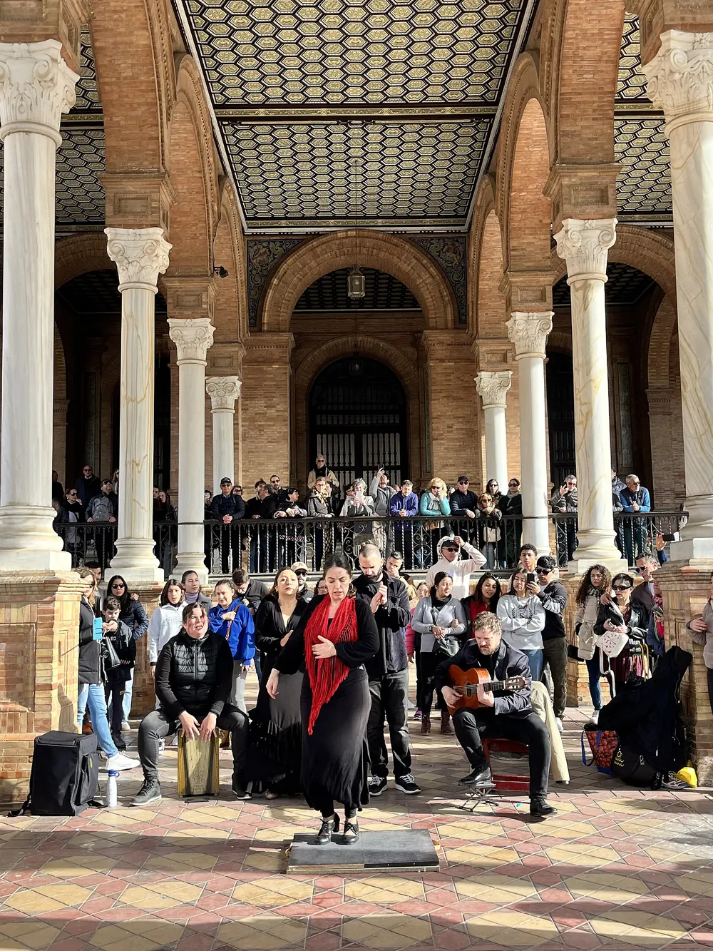A flamenco dancer in black with a red shawl beside a guitarist under the arches of the Plaza de España in Seville