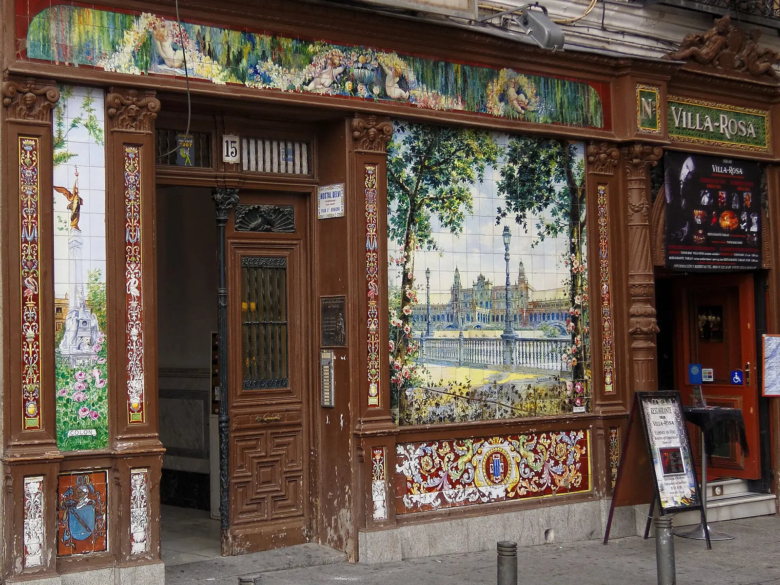 A tiled panel of a grand plaza with a lamppost beside a dark wooden door numbered 15 on the façade of the former Villa Rosa