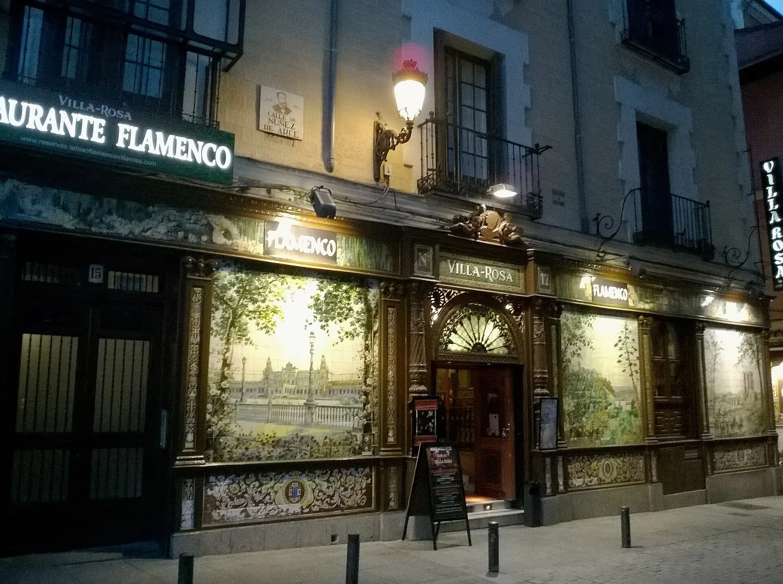 The tiled front of the former Villa Rosa on Plaza de Santa Ana at night, with lit signs and ceramic panels of Seville and other views